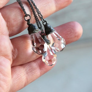 May include: Three clear crystal teardrop pendants hang from silver chains. The pendants are faceted and have a slightly iridescent quality.