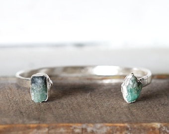 Puede incluir: Un brazalete de plata con dos piedras preciosas verdes sin pulir. El brazalete está abierto y tiene un diseño sencillo.