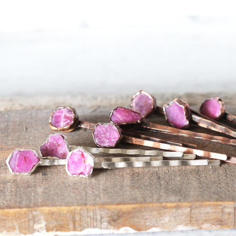 May include: A collection of hair pins with raw, pink gemstone accents. The gemstones are set in silver and copper-toned metal. The pins are arranged on a wooden surface, showcasing their natural beauty and unique design. These hair accessories are perfect for adding a touch of elegance.