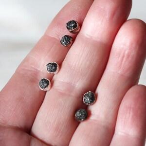 May include: Five small black diamond earrings with silver settings. The earrings are displayed on a hand.