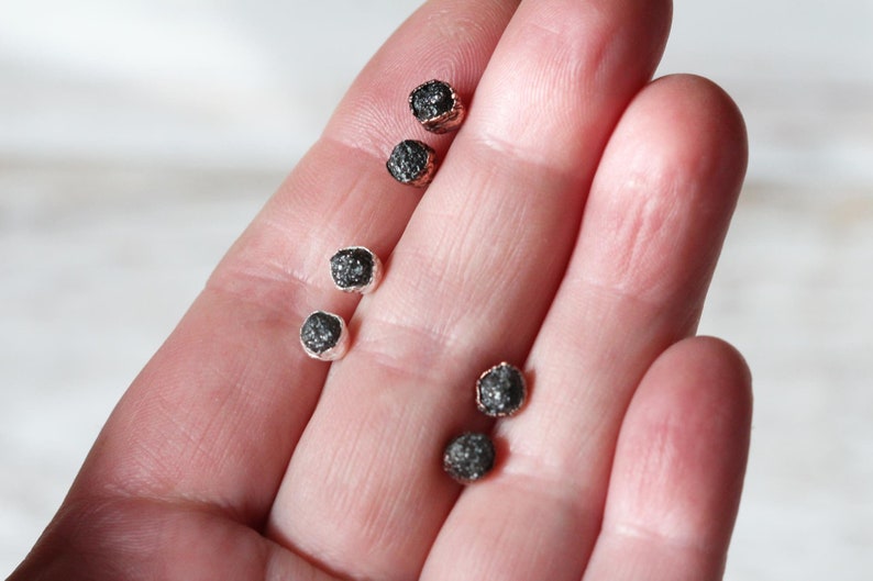 May include: Five small black diamond earrings with silver settings. The earrings are displayed on a hand.