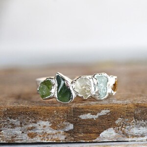May include: A silver ring with five raw gemstones set in a row. The stones are green, white, light blue, and yellow.