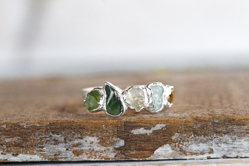 May include: A silver ring with five raw gemstones set in a row. The stones are green, white, light blue, and yellow.