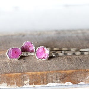 May include: Three silver-toned bobby pins with raw, vibrant pink ruby stones. The gemstones are set in a silver bezel, showcasing their natural, irregular shapes. The bobby pins rest on a weathered wooden surface.