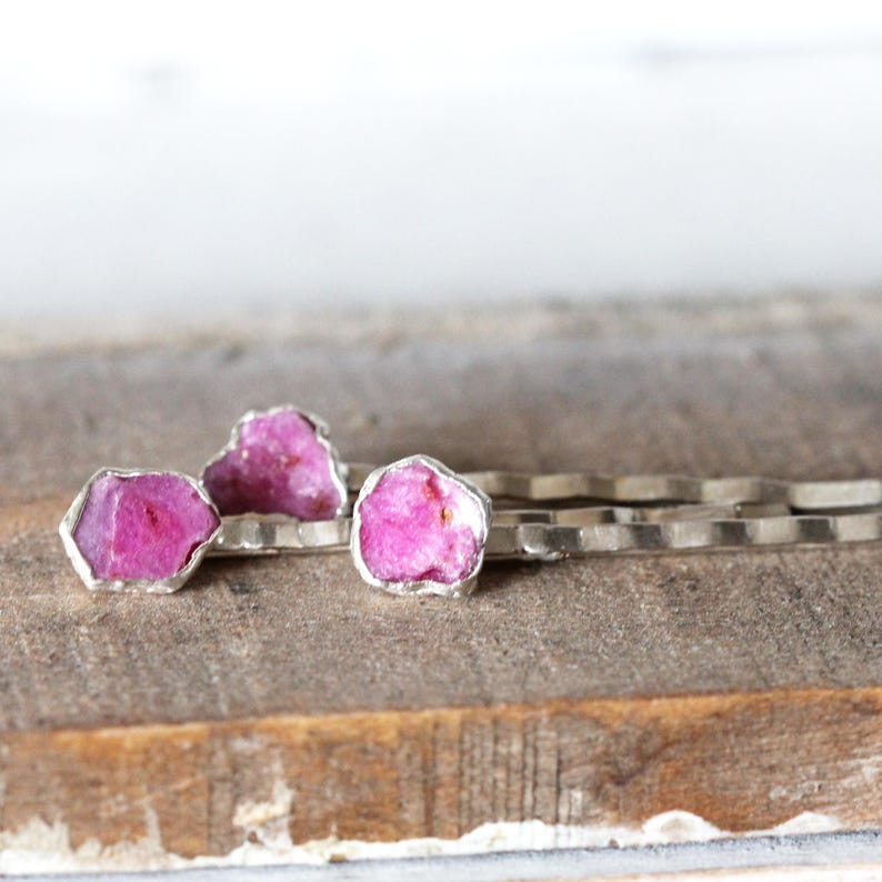 May include: Three silver-toned bobby pins with raw, vibrant pink ruby stones. The gemstones are set in a silver bezel, showcasing their natural, irregular shapes. The bobby pins rest on a weathered wooden surface.