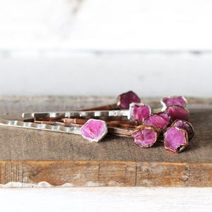 May include: A collection of hair pins with raw, pink gemstone accents. The gemstones are set in silver and gold-toned metal, creating a contrast against the natural wood surface. The pins are arranged in a casual, artistic display.