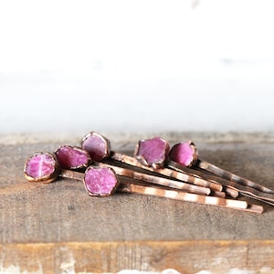 May include: A collection of copper-colored bobby pins, each adorned with a raw, vibrant pink gemstone. The gemstones are set in a delicate, gold-toned bezel, creating a contrast against the copper pins. The pins are arranged on a wooden surface.