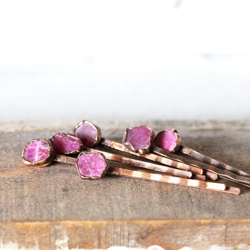 May include: A collection of copper-colored bobby pins, each adorned with a raw, vibrant pink gemstone. The gemstones are set in a delicate, gold-toned bezel, creating a contrast against the copper pins. The pins are arranged on a wooden surface.