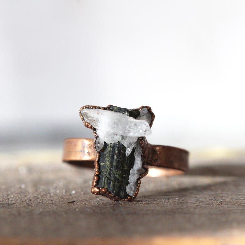 May include: A copper-colored ring featuring a dark green and white crystal centerpiece. The crystal has a rough, natural shape and is set in a copper bezel. The ring band is a simple, polished copper.
