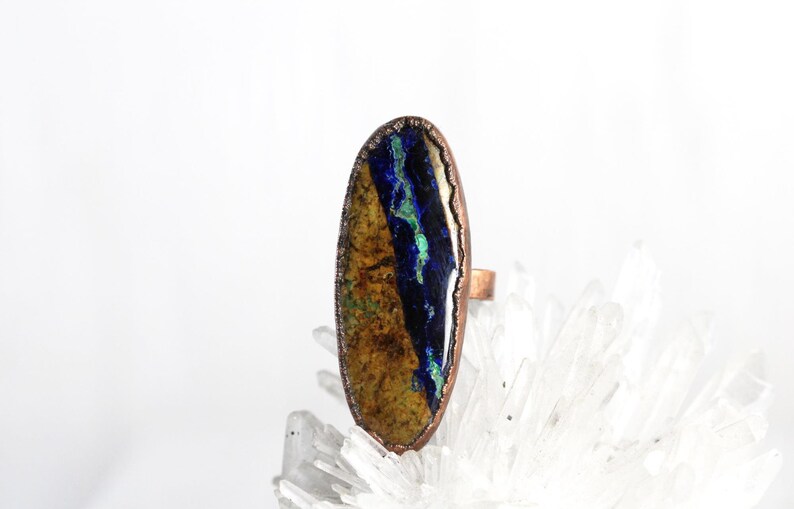 May include: A copper ring with an oval-shaped stone set in the center. The stone is a mix of blue, green, and brown colors.
