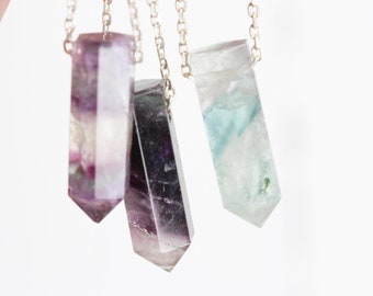 May include: Three silver necklaces with different colored fluorite crystal pendants. The pendants are all in the shape of a long, pointed rectangle. The fluorite crystals are purple, green, and a mix of purple and green.