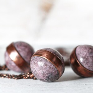 May include: Three round purple and copper pendant charms on a copper chain. The charms are made of a speckled purple stone with a copper band around the middle.