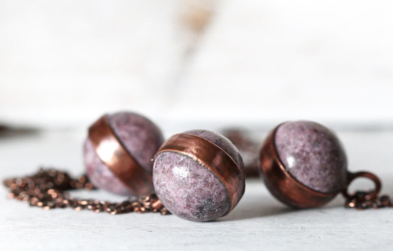 May include: Three round purple and copper pendant charms on a copper chain. The charms are made of a speckled purple stone with a copper band around the middle.