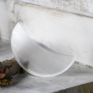 May include: A clear, crescent-shaped selenite crystal, resting on a small branch with moss.