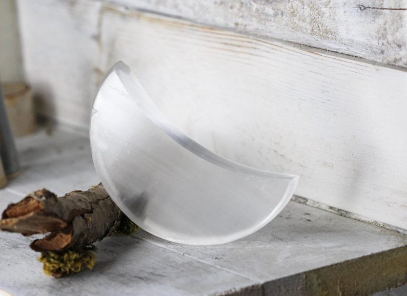 May include: A clear, crescent-shaped selenite crystal, resting on a small branch with moss.
