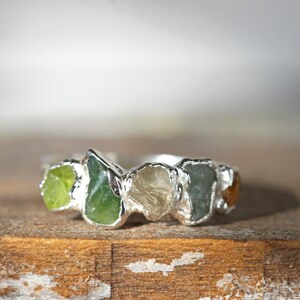 May include: A silver ring with five different colored gemstones set in the band. The gemstones are green, clear, and brown.