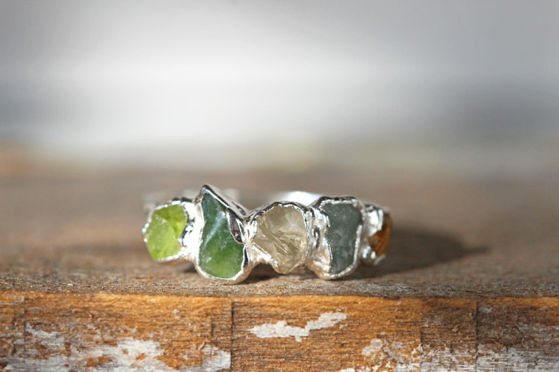 May include: A silver ring with five different colored gemstones set in the band. The gemstones are green, clear, and brown.
