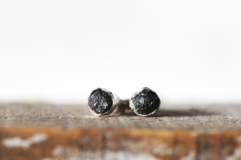 May include: A pair of silver stud earrings with black rough diamonds. The earrings are set on a wooden surface.
