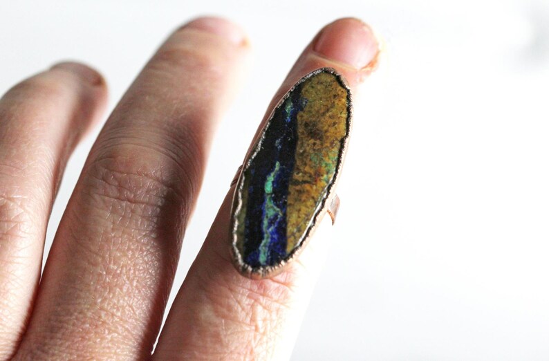 May include: A copper ring with a large oval-shaped stone set in the center. The stone is a mix of blue, green, and brown colors.