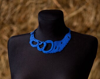May include: A vibrant blue, handmade crochet necklace displayed on a black mannequin. The necklace features a unique openwork design with circular and curved elements. The background is a blurred view of hay.