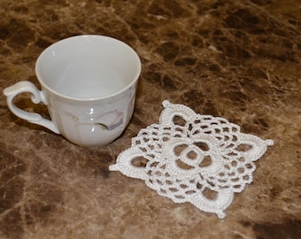May include: A white ceramic teacup with a curved handle and a delicate floral pattern, positioned next to a square, white crocheted coaster featuring an intricate, openwork lace design with a central floral motif and scalloped edges.
