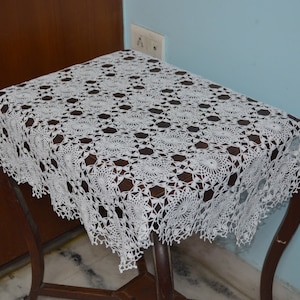 以下が含まれることがあります： 小さな暗い木製のテーブルに掛けられた、花柄の白いかぎ針編みのレースのテーブルカバー。 テーブルには湾曲した脚があり、青い壁に配置されています。 複雑なレースのデザインが見えます。