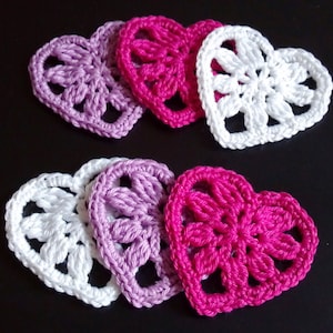 May include: A selection of handmade crochet hearts in white, light purple, and hot pink. The hearts are displayed on a dark background, highlighting their detailed openwork pattern. These decorative hearts are ideal for craft projects and embellishments.