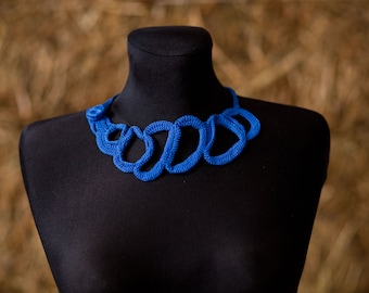 May include: A vibrant blue necklace displayed on a black mannequin. The necklace features a series of interconnected, looped designs, creating a bold and eye-catching statement piece. The background is a blurred, warm-toned texture.