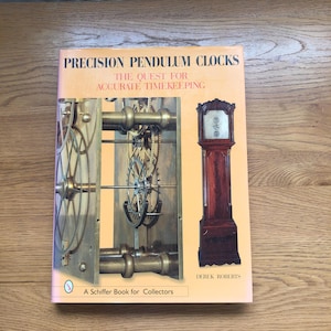 May include: A book titled "Precision Pendulum Clocks: The Quest for Accurate Timekeeping" by Derek Roberts. The cover shows clock mechanisms and a longcase clock. Published by Schiffer Book for Collectors.