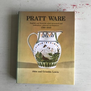 May include: A book titled "Pratt Ware" with a yellow cover and a picture of a white ceramic pitcher with a green, blue, and white floral design. The book is about English and Scottish relief decorated and underglaze colored earthenware from 1780-1840. The book is by John and Griselda Lewis.