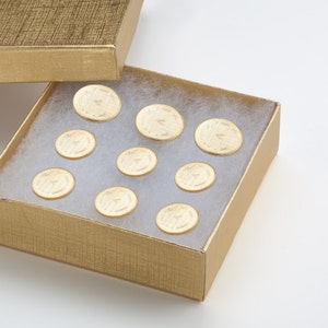 May include: A gold box with a lid open, revealing a white foam interior. Ten round, gold-colored buttons are arranged in a grid pattern inside the box.