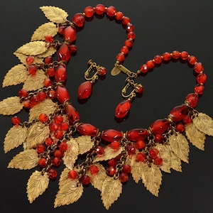 May include: A red and gold necklace and earrings set with a leaf design. The necklace features large red glass beads and gold leaf accents. The earrings are clip-on and feature a single red glass bead with a gold leaf accent.