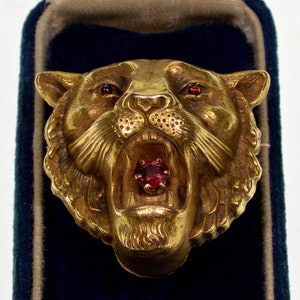 May include: A gold-toned tiger head brooch with a red gemstone in its mouth. The brooch is detailed with a textured surface and has a realistic design.