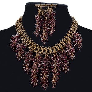 May include: A gold chain necklace with dangling clusters of purple beads. The necklace is displayed on a black mannequin bust.