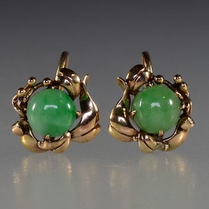 Könnte beinhalten: Ein Paar goldfarbene Ohrringe, jeweils mit einer leuchtend grünen Jadekugel. Der Jade wird von kunstvollen goldenen Metallarbeiten mit Blatt- und Perlendetails gehalten. Die Ohrringe haben einen Klappverschluss.