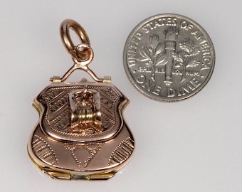Puede incluir: Un colgante dorado en forma de escudo con intrincados patrones grabados y un pequeño lazo para una cadena. Una moneda está al lado del colgante para comparar el tamaño. El colgante tiene una abertura con bisagras y un cierre decorativo.