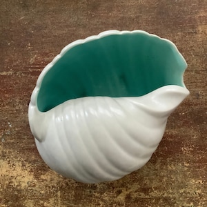 May include: A white ceramic seashell planter with a teal green interior. The planter is shaped like a seashell and has a smooth, glossy finish.