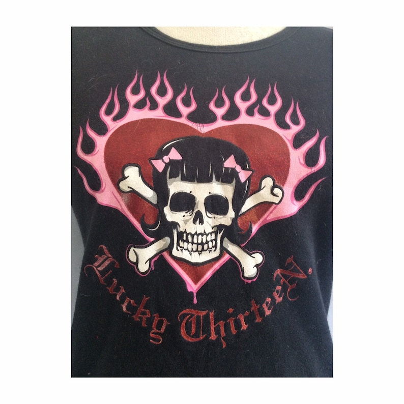 May include: Black t-shirt with a pink and red heart design featuring a skull with pink bows and crossbones. The text "Lucky Thirteen" is written in a gothic font below the heart.