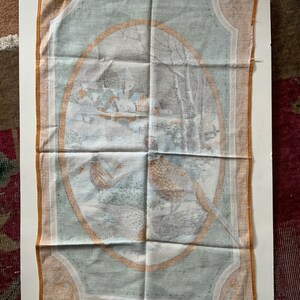 May include: A rectangular, vintage-style tea towel with a winter scene and pheasant illustrations. The central oval design features a snowy village and bare trees. The border is a light orange color.