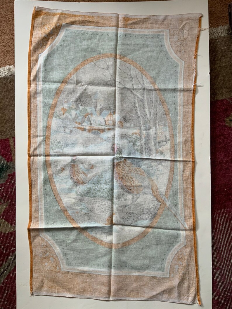 May include: A rectangular, vintage-style tea towel with a winter scene and pheasant illustrations. The central oval design features a snowy village and bare trees. The border is a light orange color.