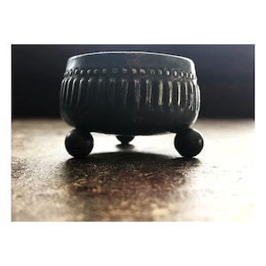 May include: A small, black, metal salt cellar with a decorative rim and three round legs.