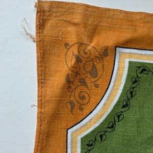 May include: Close-up of a section of a decorative textile. The design features a mustard yellow border with a corner design of swirling brown shapes. The interior has a green field with a black leaf pattern along the edge.