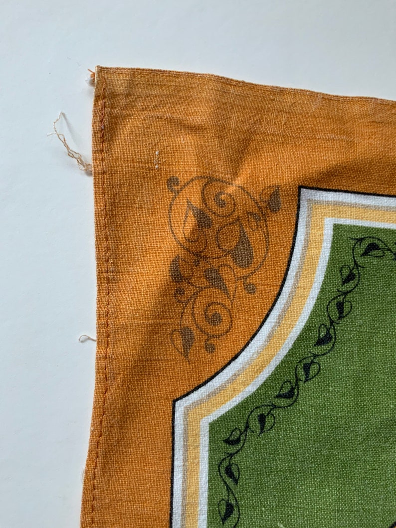 May include: Close-up of a section of a decorative textile. The design features a mustard yellow border with a corner design of swirling brown shapes. The interior has a green field with a black leaf pattern along the edge.