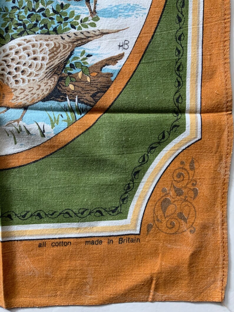 May include: A vintage cotton tea towel with a detailed illustration of a bird in a natural setting. The design is framed by a green border with decorative elements and the text "all cotton made in Britain".
