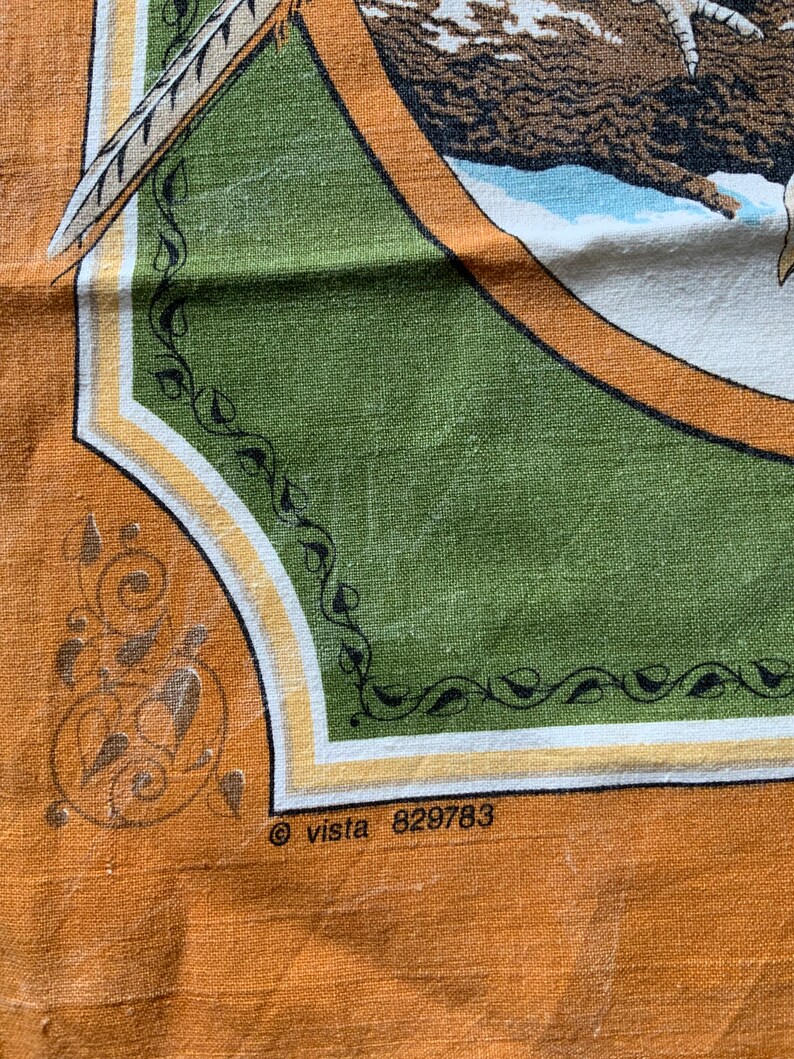 May include: Close-up of a vintage fabric with a nature-themed design. The fabric features a central green panel with a circular illustration of a bird and feather. The border is orange with decorative elements and the text "&copy; vista 829783".