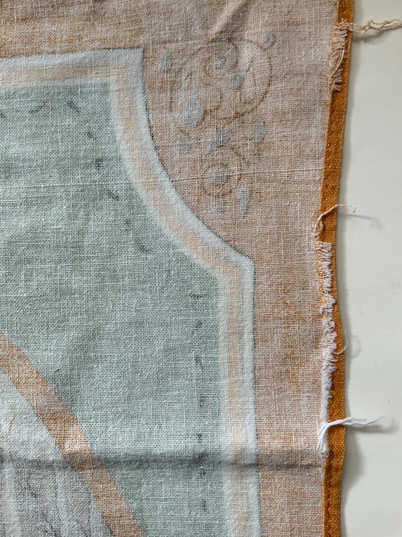 May include: Close-up of a section of fabric with a detailed pattern. The design features a combination of light green, white, and tan colors. The fabric has a textured appearance, with a frayed edge on the right side.