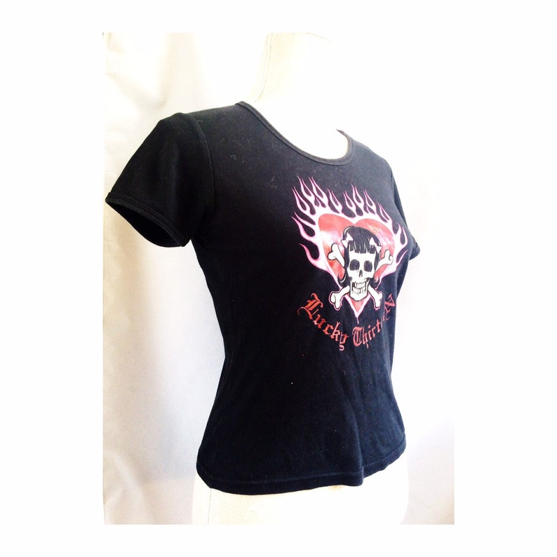 May include: Black t-shirt with a pink and red graphic of a skull with crossed bones inside a heart with flames. The text "Lucky Thirteen" is below the graphic.