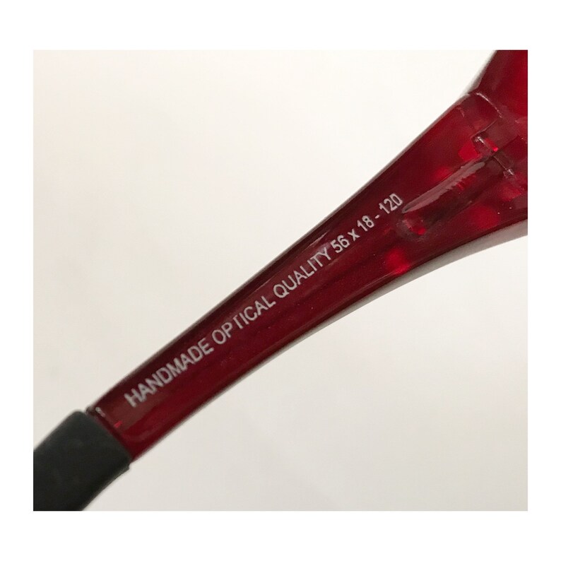 以下が含まれることがあります： 「HANDMADE OPTICAL QUALITY 56 x 18 - 120」と印刷された赤い眼鏡の腕のクローズアップ。