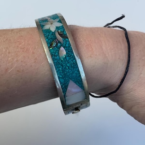 Peut inclure: Un bracelet jonc en argent avec un motif d'incrustation en turquoise et nacre. Le motif représente une fleur et des feuilles.