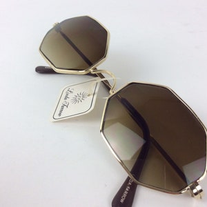 May include: Gold-rimmed sunglasses with brown lenses in an octagonal shape. The sunglasses have a brown label that reads "Linda Farrow".